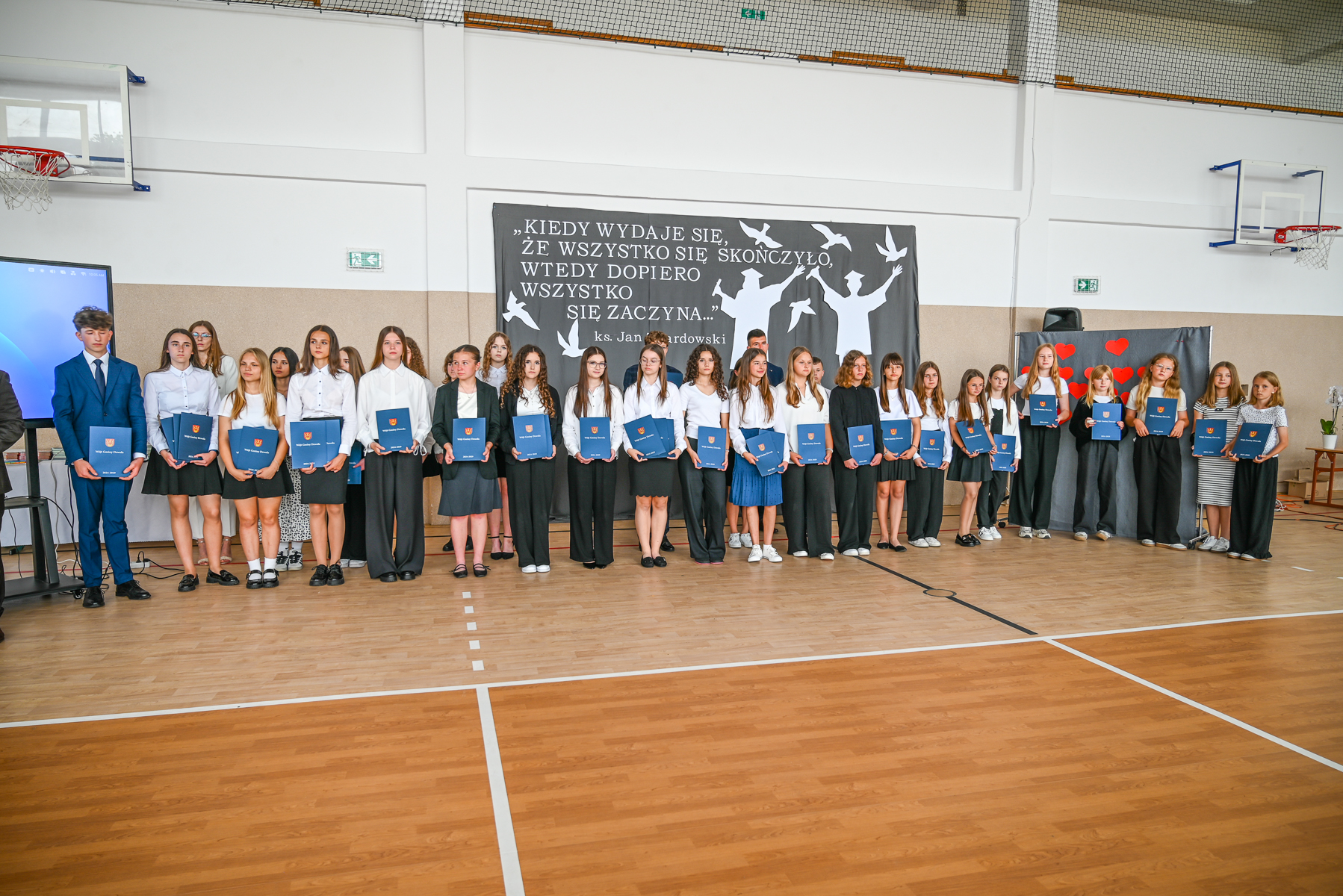 Grupa młodzieży ubranej formalnie, stojąca w rzędzie na sali gimnastycznej, trzymająca teczki, za nimi czarna plansza z białym napisem i sylwetkami ptaków i postaci.