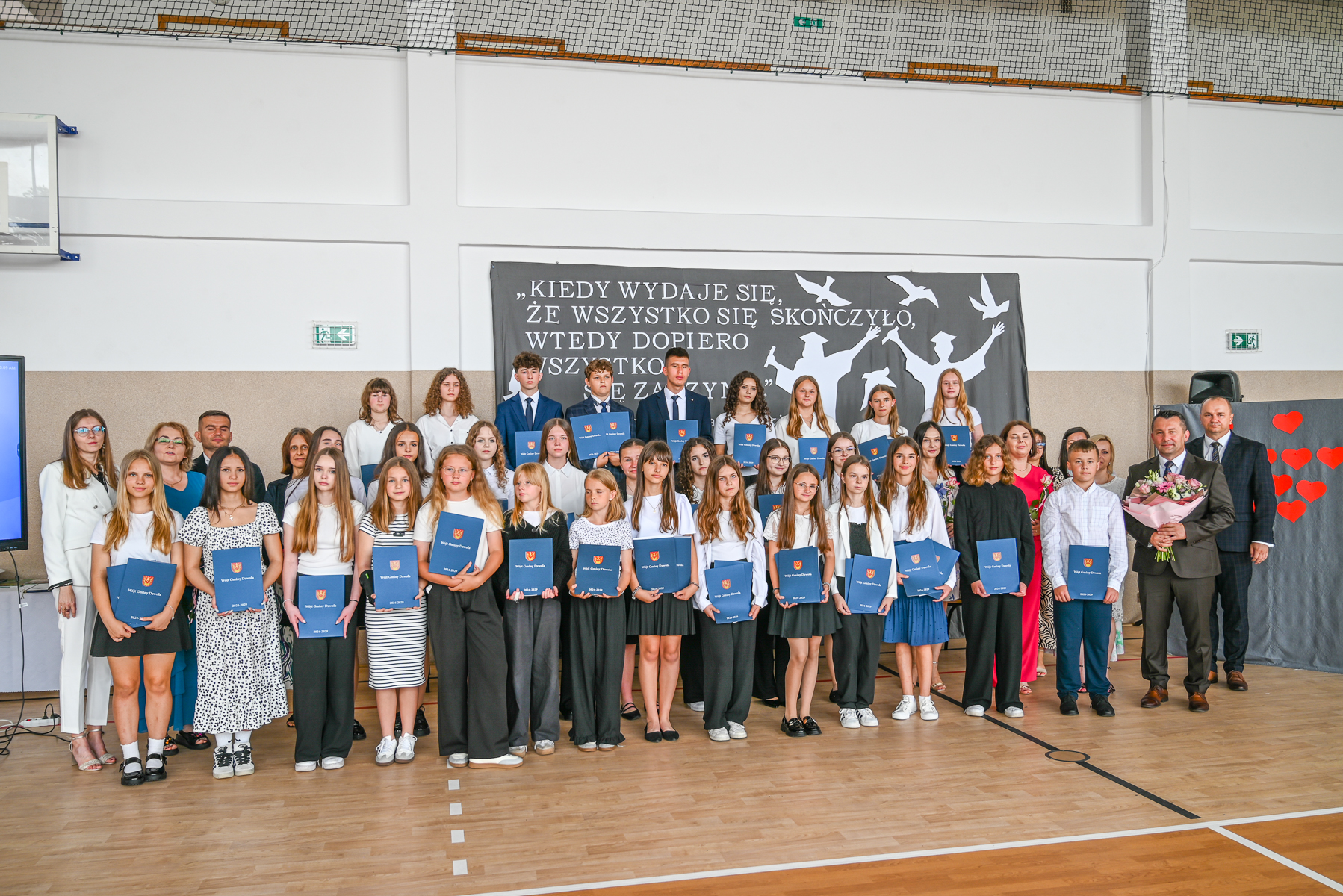 Grupa dzieci i dorosłych w sali gimnastycznej, trzymających niebieskie teczki, w tle czarne płótno z białym napisem i sylwetkami absolwentów w czapkach, drewniana podłoga.