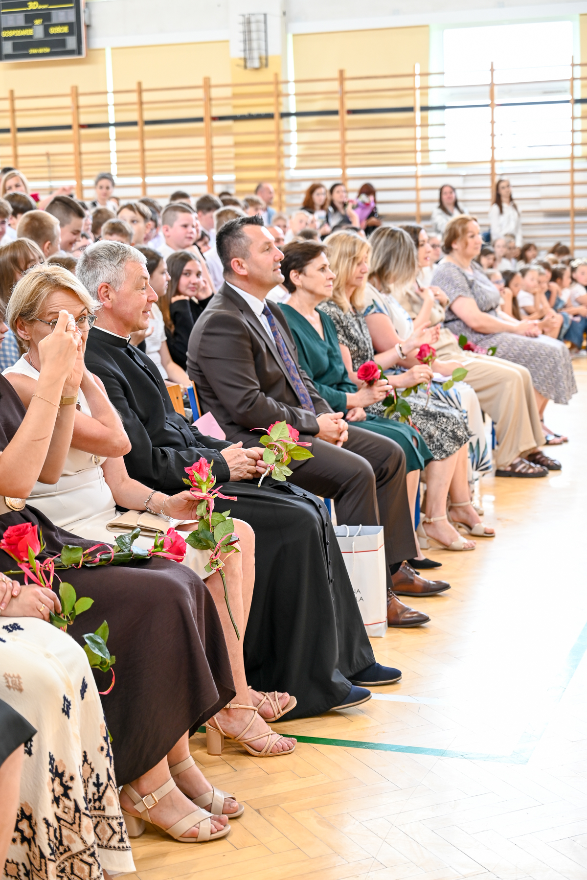Ludzie siedzący w rzędzie na drewnianej podłodze, trzymający czerwone róże, w tle dzieci i siatka gimnastyczna na ścianie hali sportowej.