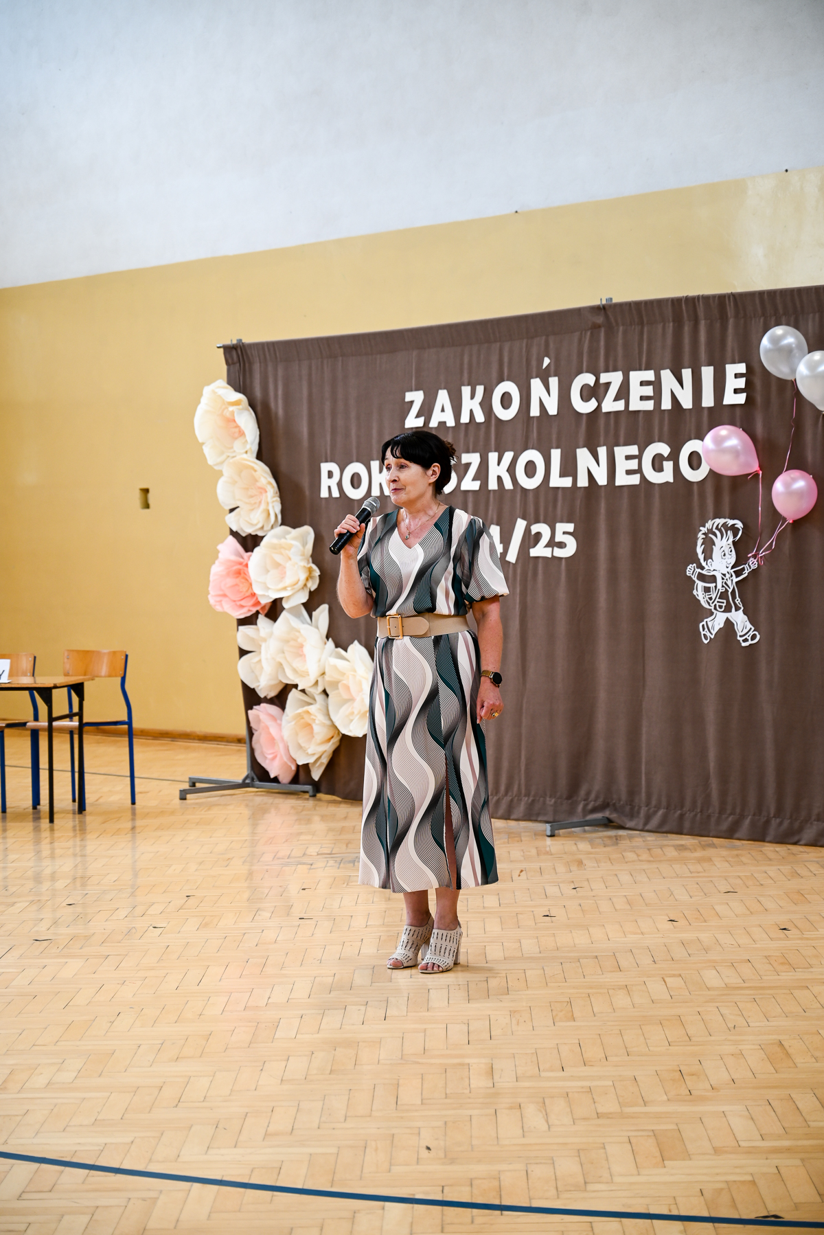 Kobieta w sukience z wzorem stojąca na parkiecie sali gimnastycznej, trzyma mikrofon, za nią brązowa kurtyna z napisem i dekoracje z balonów i kwiatów z papieru.