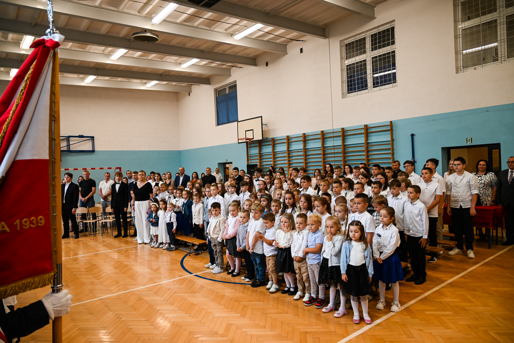 Grupa dzieci i dorosłych ubranych na biało i czarno stoi w rzędach na sali gimnastycznej z drewnianą podłogą i niebiesko-białymi ścianami, obok widoczna czerwona flaga.