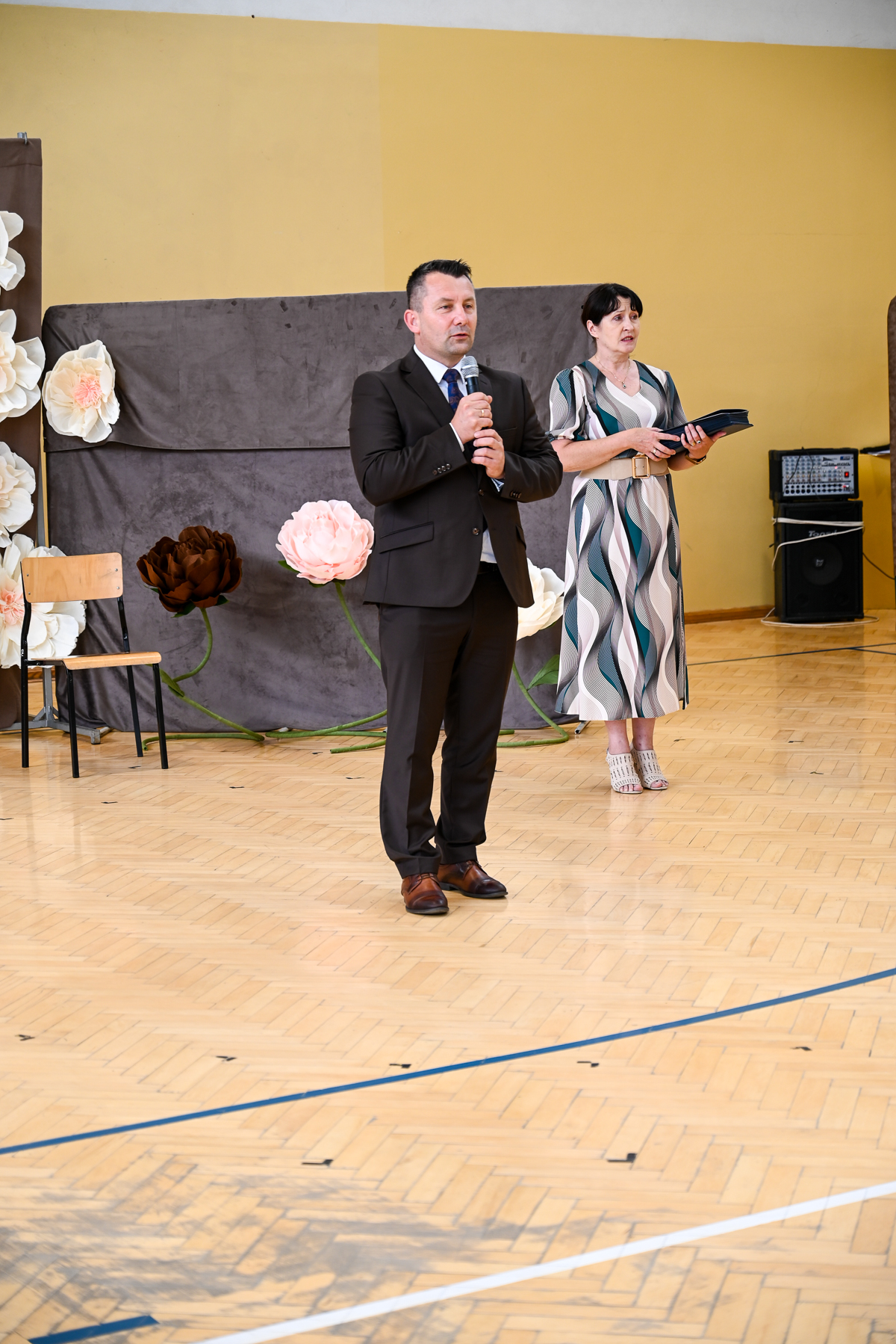 Mężczyzna w ciemnym garniturze z mikrofonem i kobieta w wzorzystej sukience na drewnianej sali z dekoracją kwiatową i nagłośnieniem.