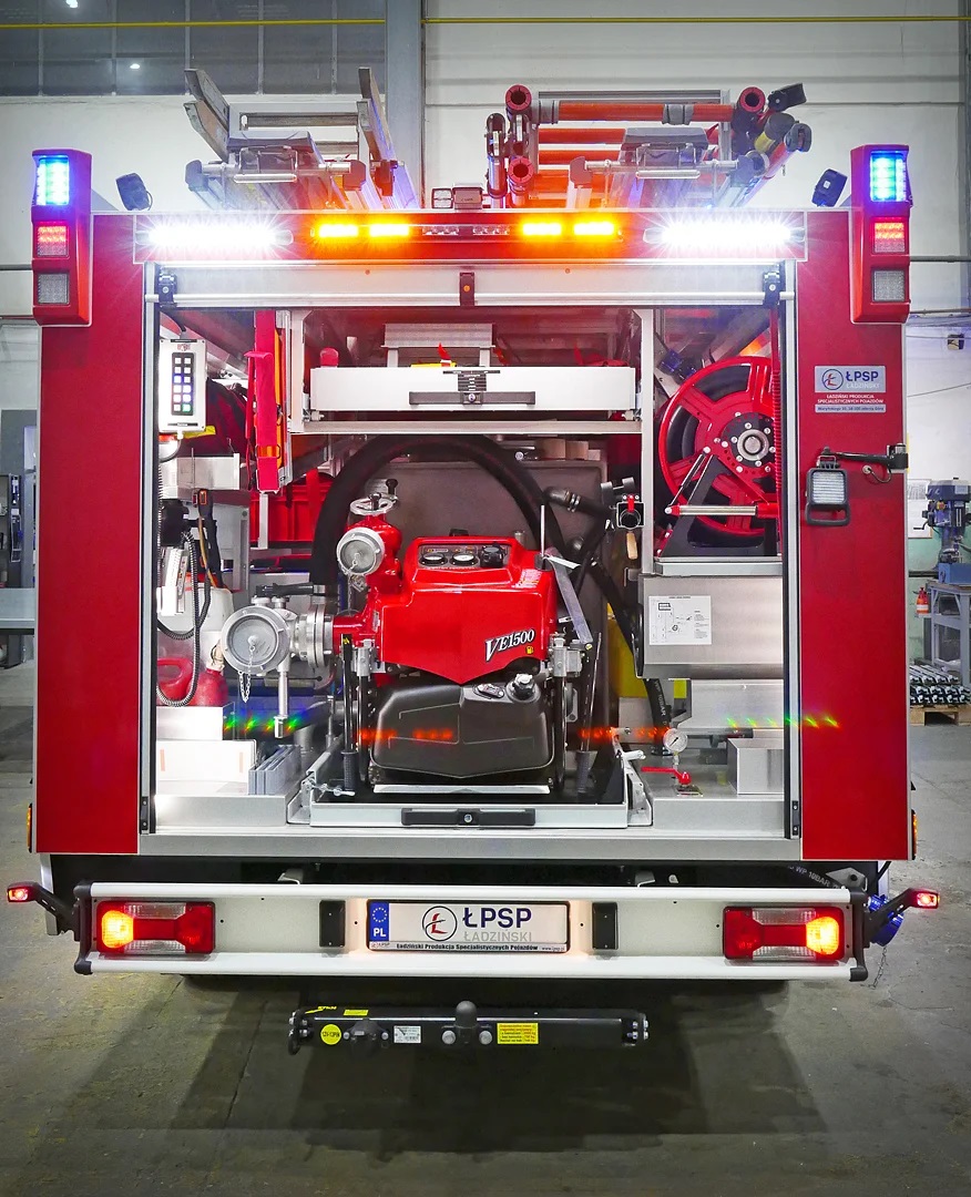 Czerwony sprzęt pożarniczy z wężem, kołem i pompą, światła ostrzegawcze na górze, tablica rejestracyjna, wnętrze hali przemysłowej.