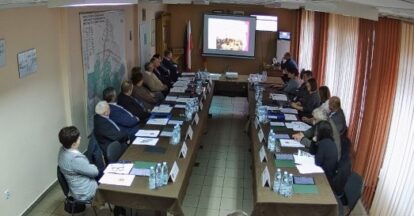 Sala konferencyjna z dwoma rzędami stołów ustawionymi w podkowę, kilkanaście osób siedzących, projektor z wyświetlanym obrazem, butelki wody na stołach.