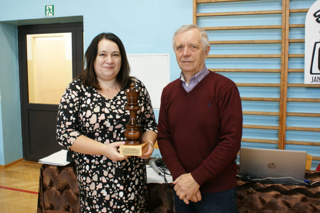 Kobieta w czarnej sukience w jasne plamy trzyma dużą drewnianą figurę szachową, obok stoi mężczyzna w bordowym swetrze, w tle niebieska ściana i laptop na stole.