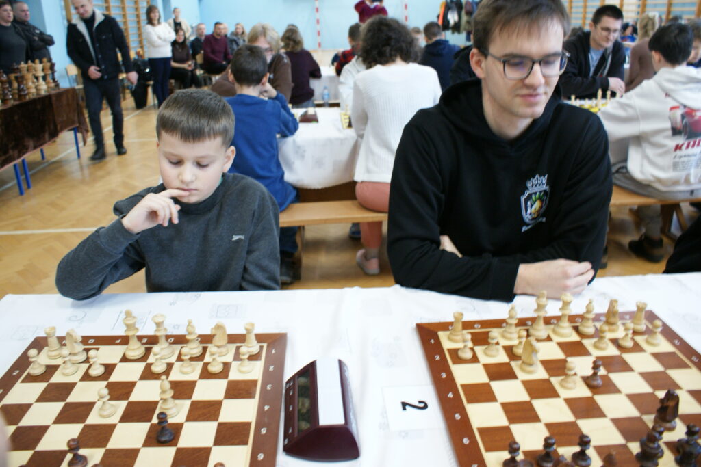 Dwóch graczy przy szachownicach na stole, chłopiec w szarej bluzie, mężczyzna w czarnej bluzie z kapturem i okularami, w tle inni uczestnicy i widzowie w sali sportowej.