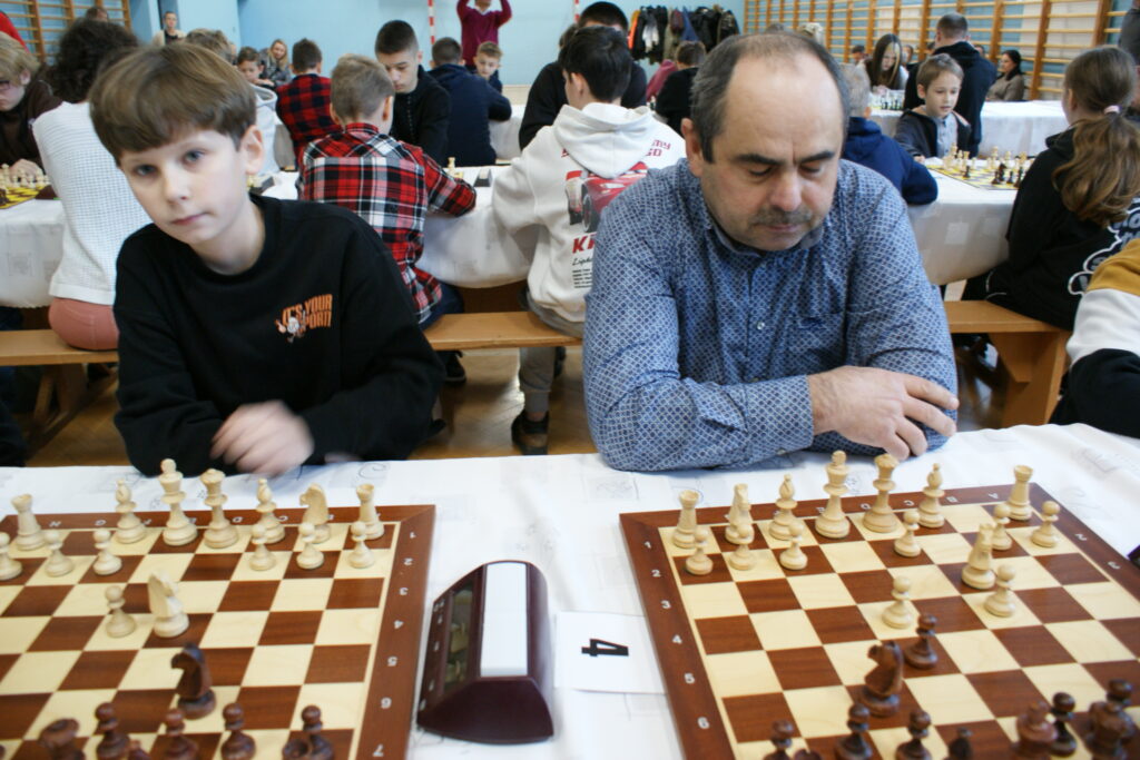 Dwaj mężczyźni grający w szachy przy stole z zegarem, w tle inne osoby siedzące na ławkach i grające w szachy, jasna sala z niebieskimi ścianami i drabinkami gimnastycznymi.