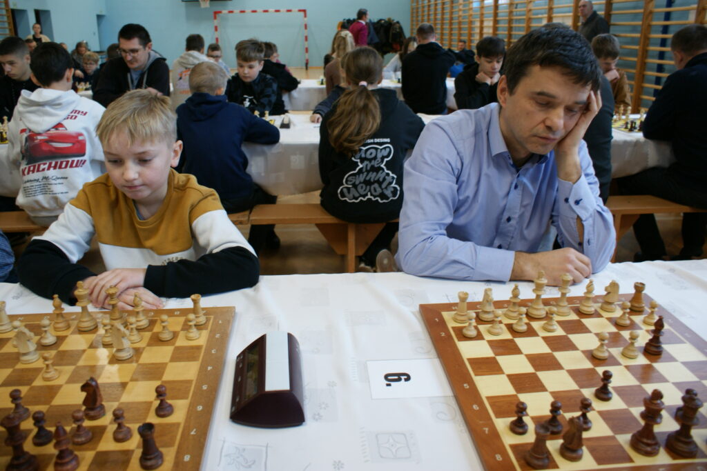 Dziecko w bluzie w kolorach białym, czarnym i żółtym oraz mężczyzna w jasnoniebieskiej koszuli siedzą przy dwóch szachownicach na stole z białym obrusem w sali z drewnianymi ławkami i ścianką gimnastyczną.