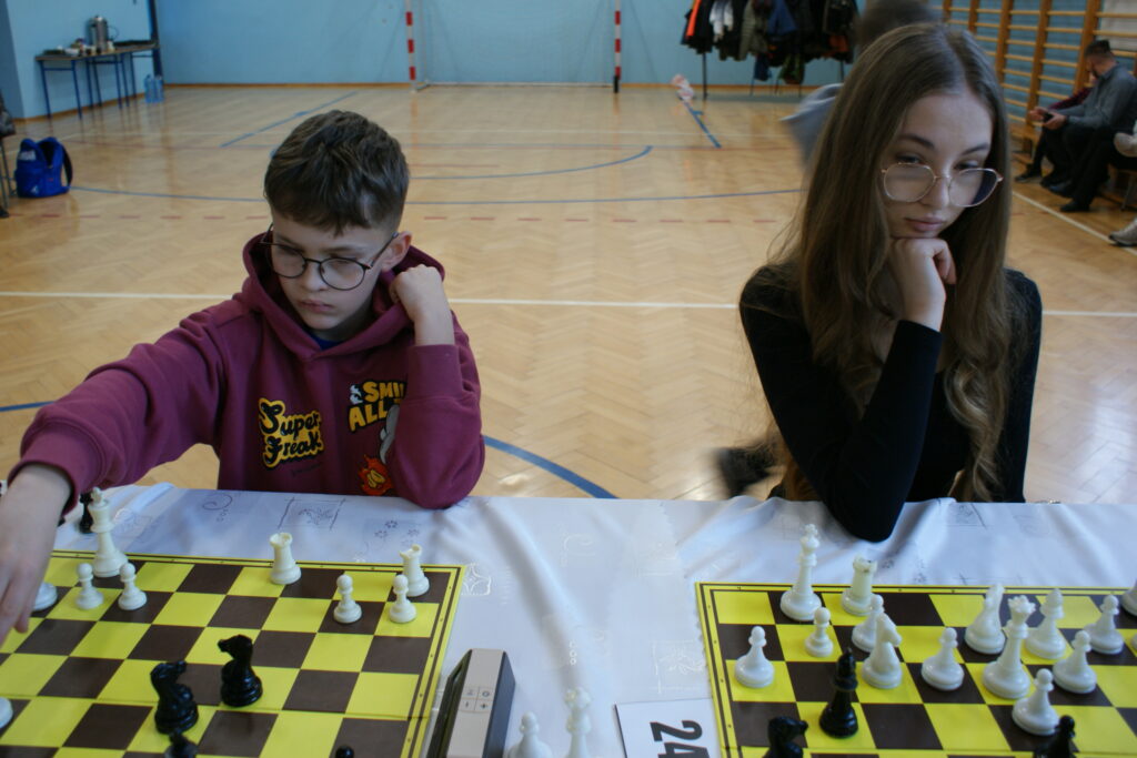 Chłopiec w bordowej bluzie i dziewczyna w czarnym ubraniu grają w szachy przy dwóch żółto-brązowych planszach na stole z białym obrusem w sali gimnastycznej.