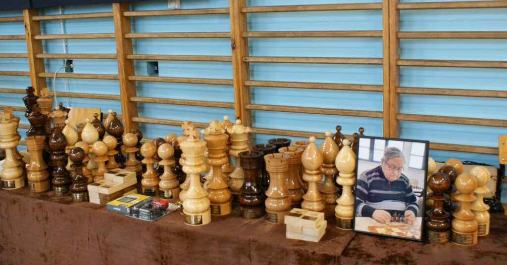 Duże drewniane figury szachowe w jasnym i ciemnym kolorze ułożone na stole z brązowym obrusem, za nimi ściana z drewnianymi poręczami, na stole zdjęcie starszego mężczyzny grającego w szachy.