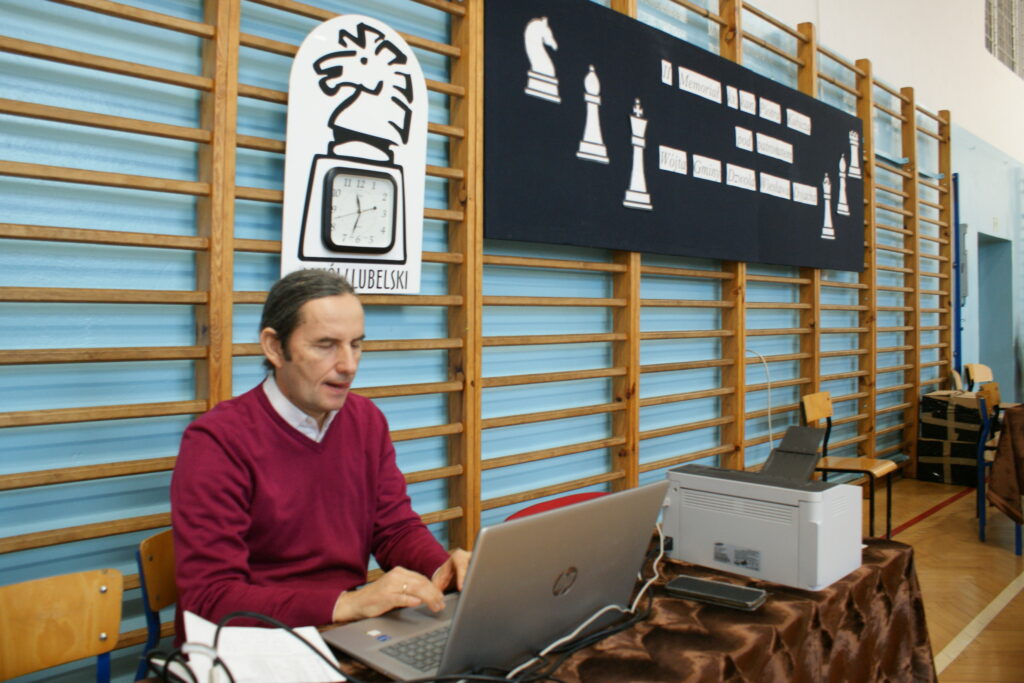 Mężczyzna w bordowym swetrze pracuje na laptopie przy stole z brązowym obrusem, obok drukarka, w tle niebieska ściana z drabinkami i plansza z szachowymi figurami.