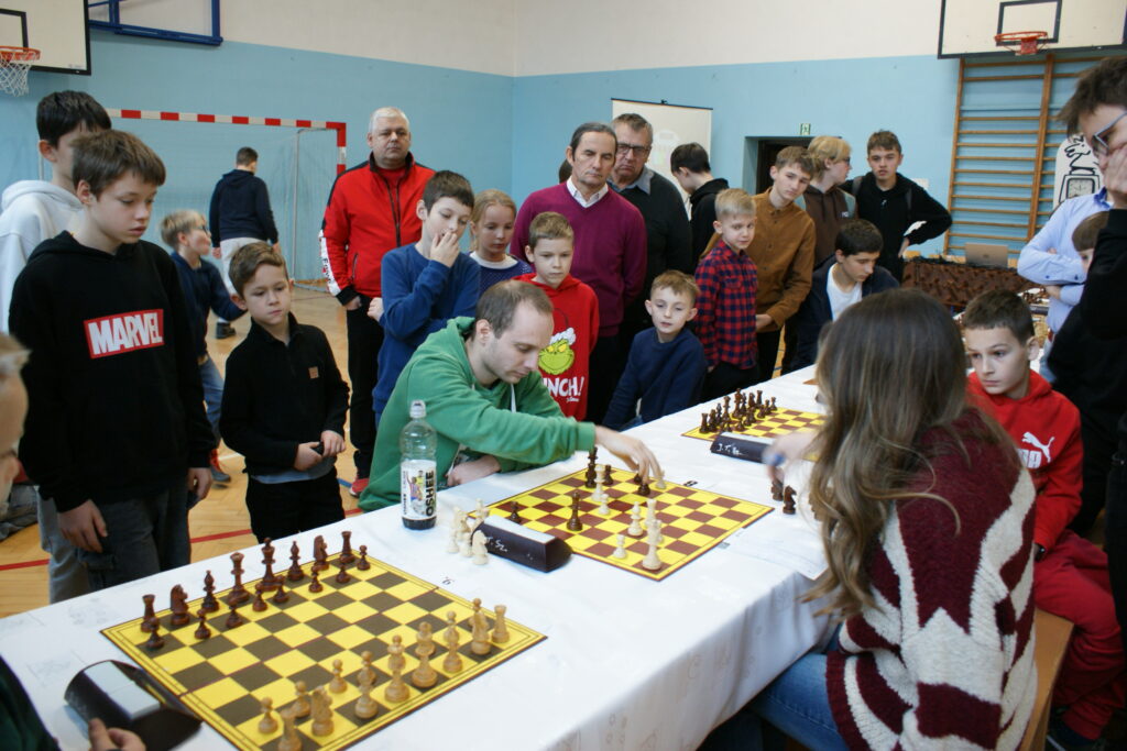 Grupa dzieci i dorosłych obserwuje grę w szachy przy stołach z białymi obrusami w sali gimnastycznej z niebieskimi ścianami i parkietem.
