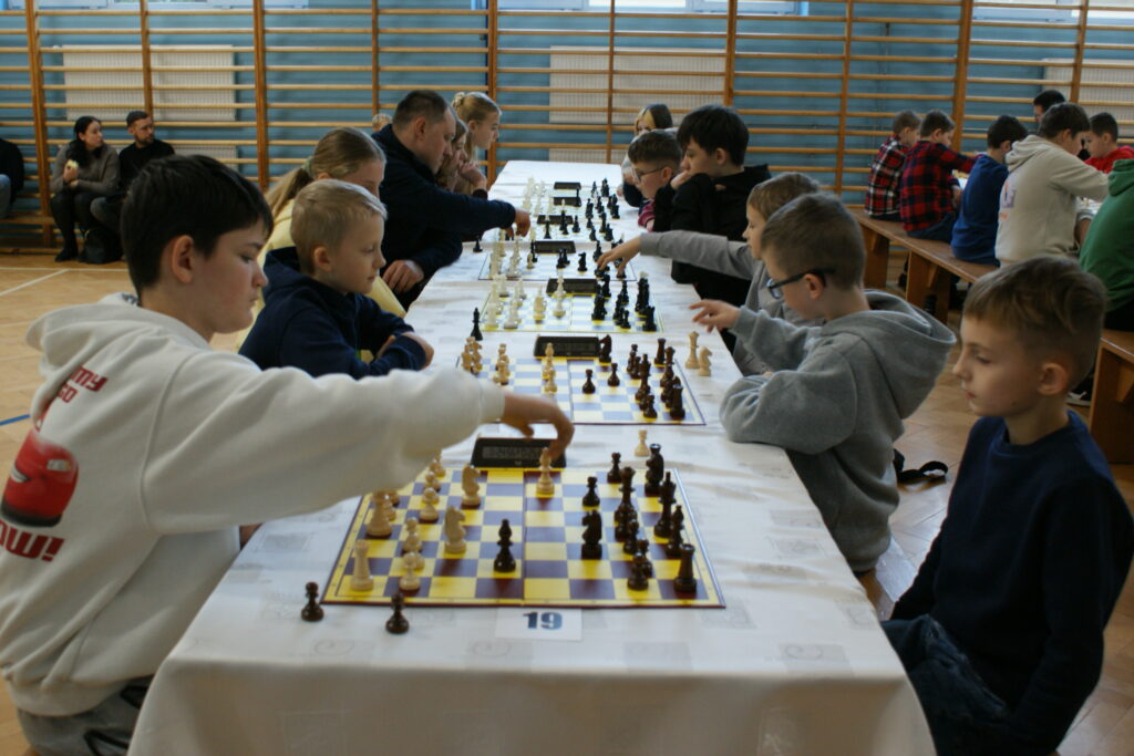 Dzieci i dorośli grający w szachy przy długim białym stole, plansze z ciemno-żółtymi polami, drewniane ściany sali gimnastycznej, jasna podłoga.