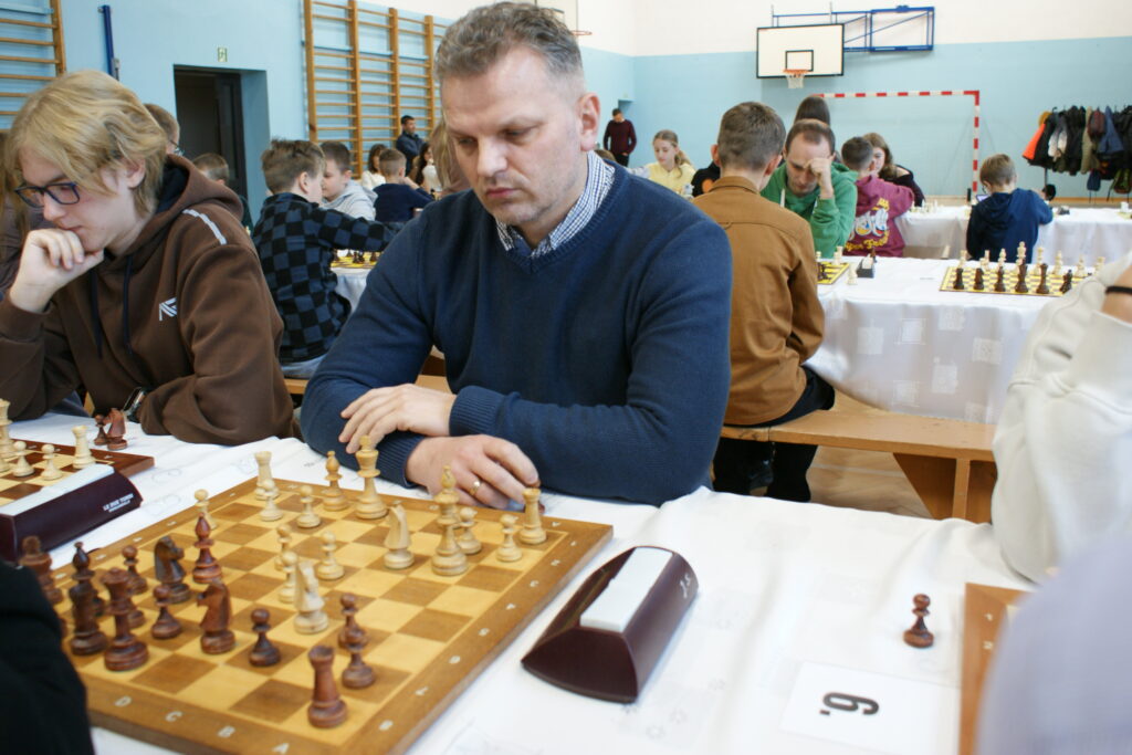 Mężczyzna w niebieskim swetrze gra w szachy przy stole z białym obrusem, w tle inne osoby grające w szachy w sali gimnastycznej z niebieskimi ścianami.