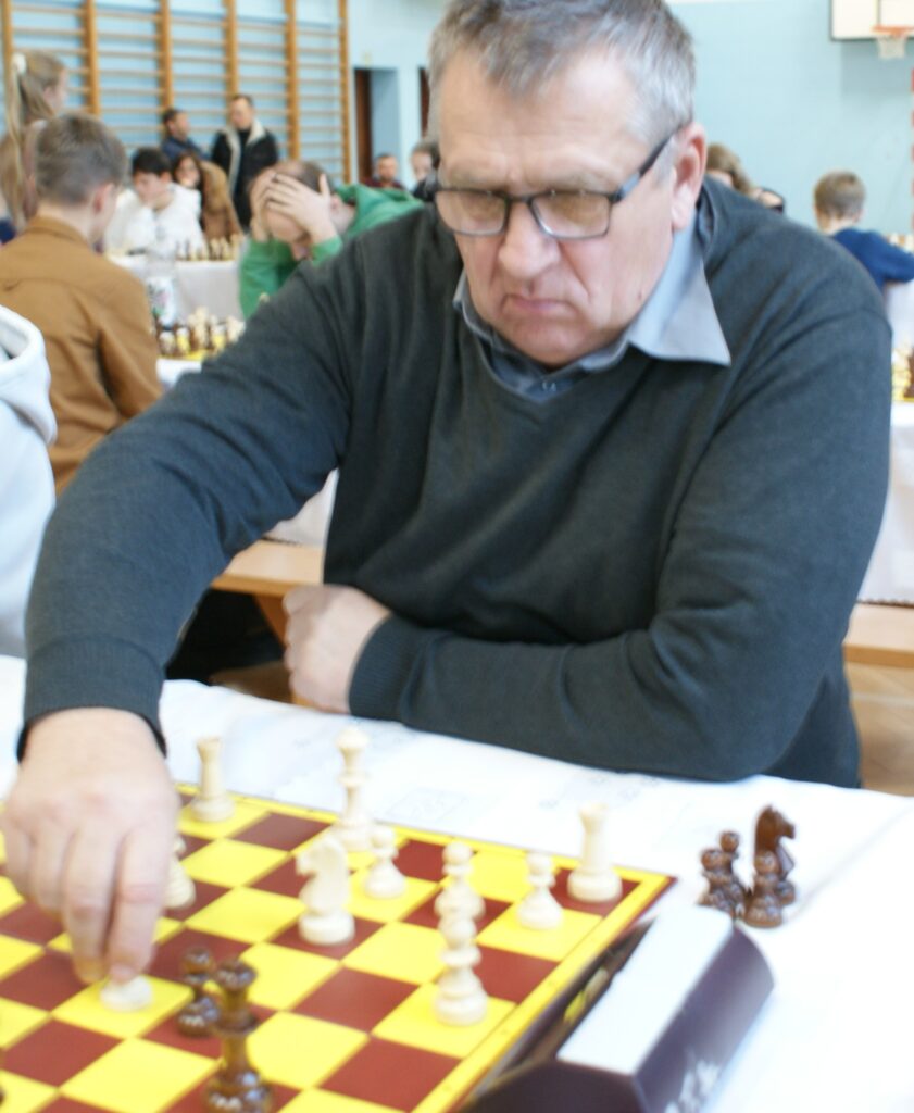 Mężczyzna w szarym swetrze przesuwa białą figurę na żółto-brązowej planszy do szachów, w tle inni grający i drewniane drabinki na ścianie.