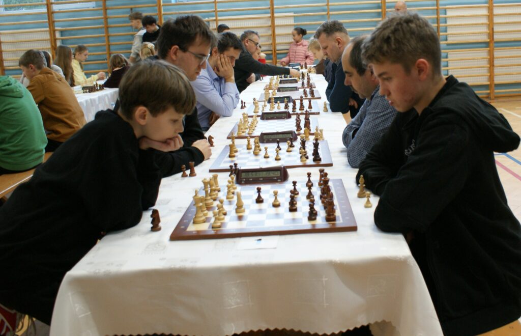 Kilka par osób siedzi po obu stronach stołu z białym obrusem, grając w szachy na drewnianych planszach w dużej sali z niebieskimi ścianami i drabinkami gimnastycznymi.