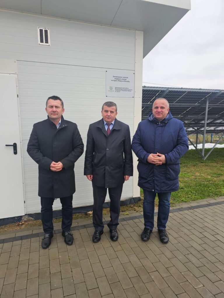 Trzej mężczyźni w ciemnych płaszczach stoją na szarej kostce brukowej przed jasnoszarym budynkiem z tablicą, obok metalowa konstrukcja paneli słonecznych na tle trawy i nieba.