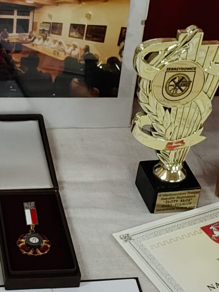 Złoty puchar z czarną podstawą, medalik w etui z ciemnym wnętrzem, częściowo widoczny dyplom z herbem, na tle zdjęcie ludzi siedzących przy stole.