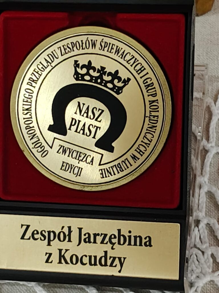 Złota okrągła plakieta z czarnym herbem i napisem na czerwonym tle oraz złota tabliczka z czarnym napisem "Zespół Jarzębina z Kocudzy".