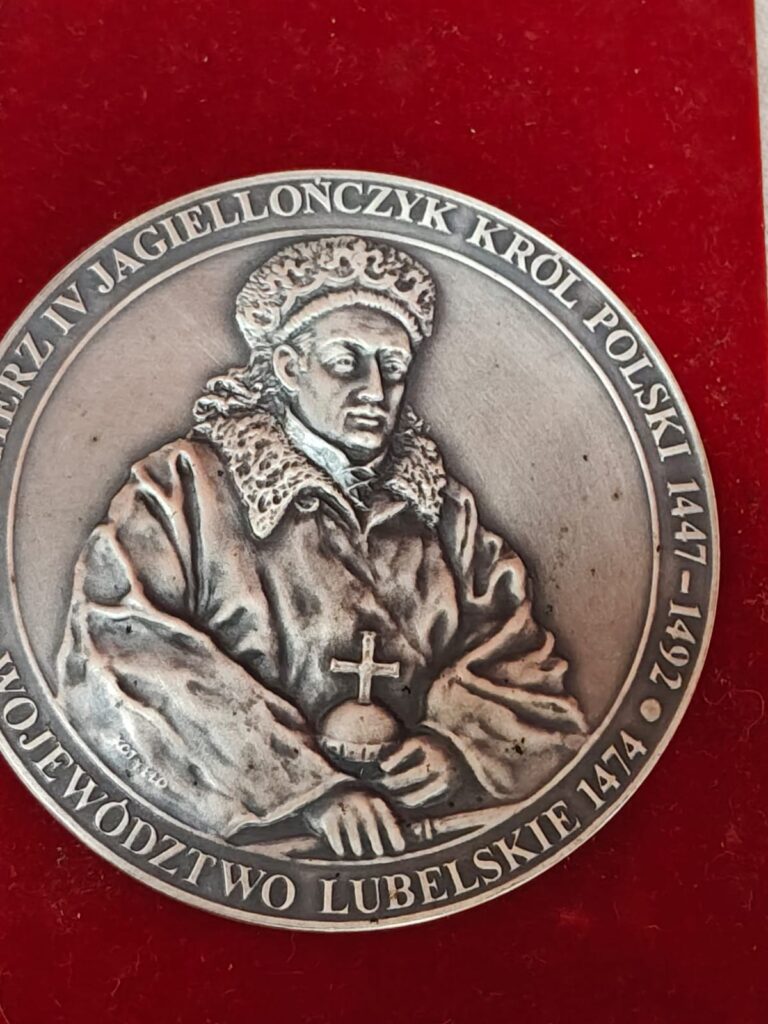 Moneta lub medal z wizerunkiem mężczyzny w koronie i futrzanym płaszczu, trzymającego kulę z krzyżem, na czerwonym tle, z napisem wokół postaci.