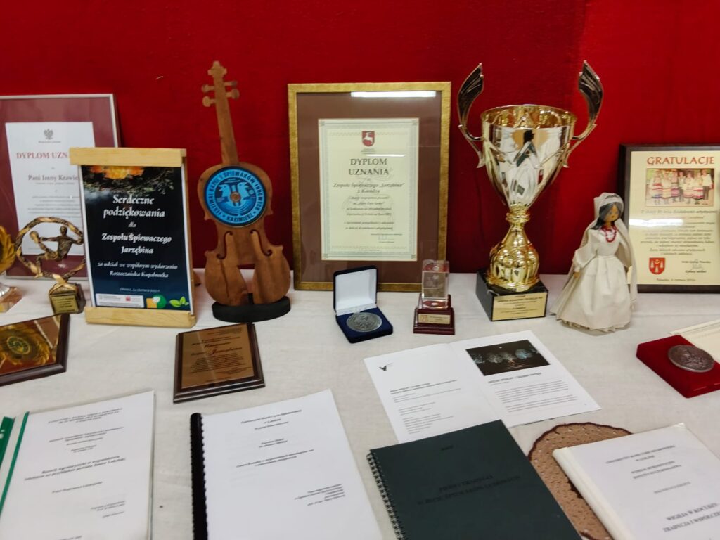 Stół z dokumentami, dyplomami, medalami, złotym pucharem, drewnianą dekoracją w kształcie skrzypiec i lalką w białej tradycyjnej sukni na czerwonym tle.