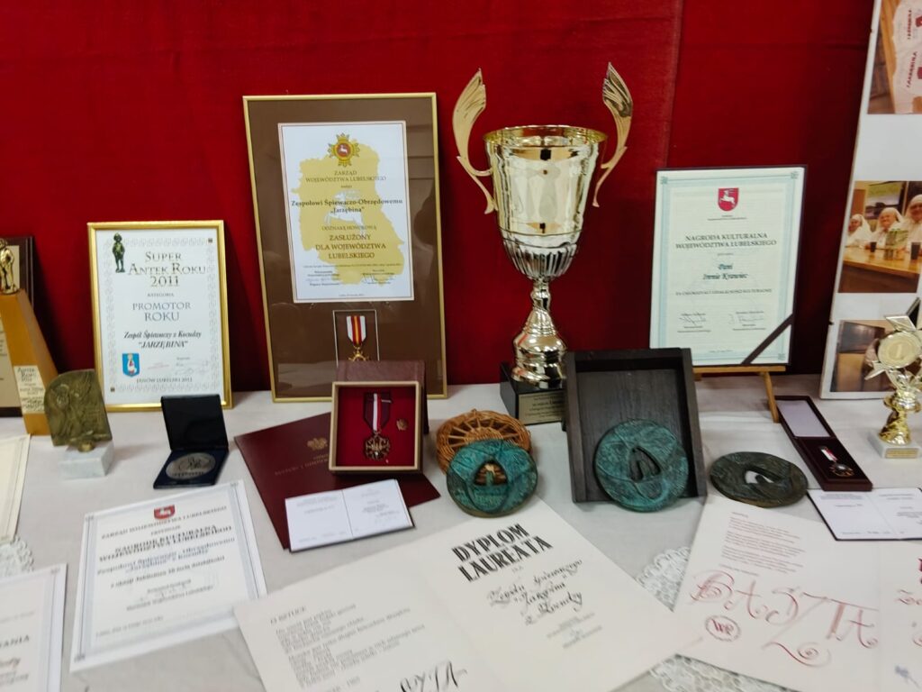 Stół z certyfikatami, dyplomami, medalami, pucharem i statuetkami na białym obrusie na tle czerwonej ściany.