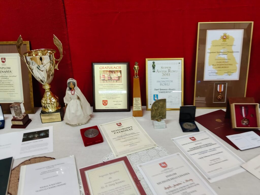 Złoty puchar, lalka w białej sukni, dyplomy, medale i statuetki na białym stole na tle czerwonej tkaniny.