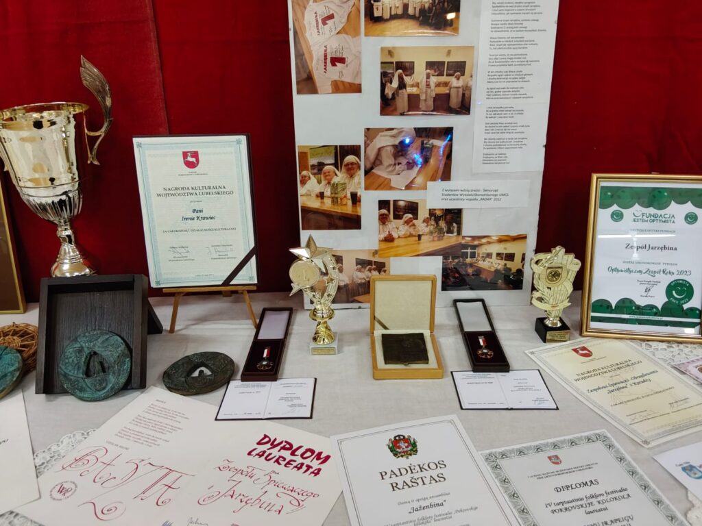 Stół z nagrodami i dyplomami, puchary srebrny i złoty, medale, plakaty ze zdjęciami osób w białych strojach, czerwone tło.