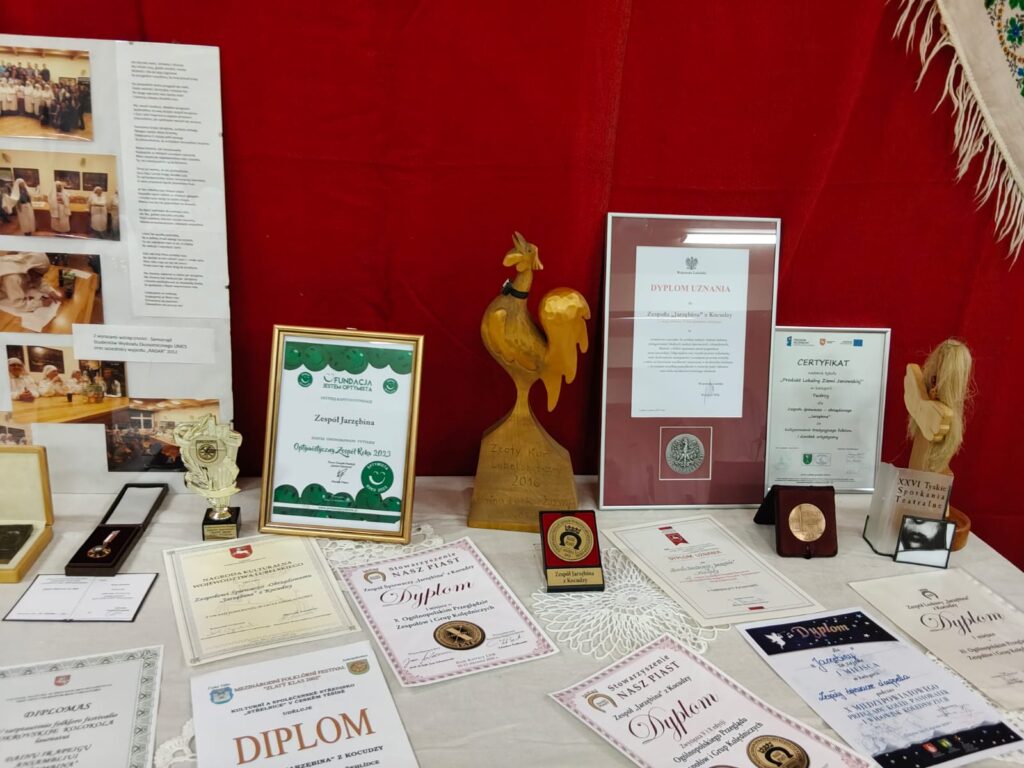 Stół z białym obrusem, na nim liczne dyplomy, certyfikaty, medale i statuetki na tle czerwonej tkaniny. Drewniana statuetka koguta i figurka anioła.