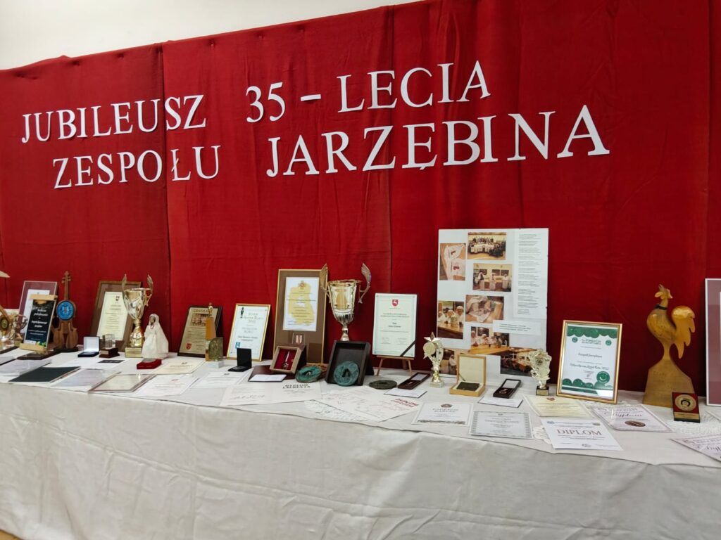 Napis na czerwonym tle "Jubileusz 35-lecia zespołu Jarzębina", pod nim stół z licznymi pucharami, dyplomami, medalami i trofeami.