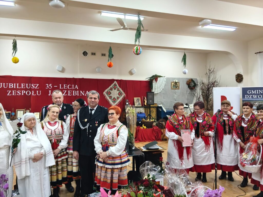 Grupa osób w tradycyjnych strojach ludowych i mundurach stojących na tle czerwonej dekoracji z napisem, w sali z wiszącymi kolorowymi ozdobami i kwiatami na pierwszym planie.