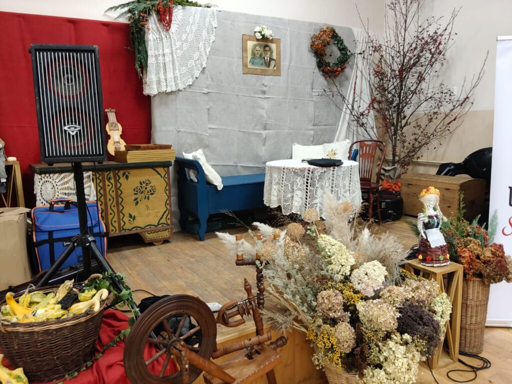 Wnętrze z drewnianą podłogą, koszami z suszonymi kwiatami, obrusem szydełkowym na okrągłym stole, niebieską ławką, starym meblem, ramką ze zdjęciem, dużym głośnikiem i kołowrotkiem.