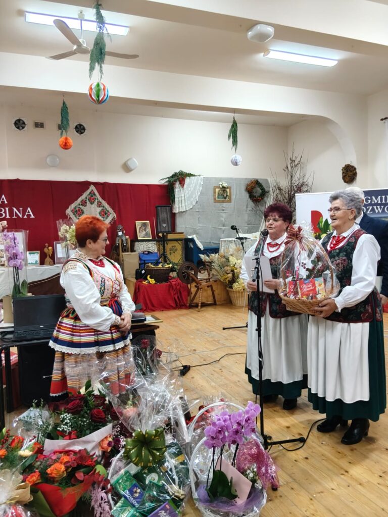 Trzy starsze kobiety w tradycyjnych strojach ludowych, dwie trzymają kosz prezentowy, w tle dekoracje ścienne i kwiaty w opakowaniach na podłodze.