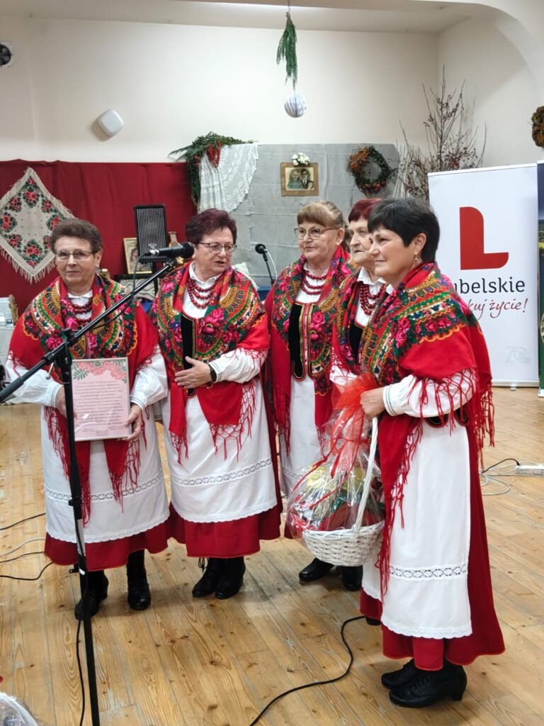Pięć kobiet w tradycyjnych białych sukienkach z czerwonymi spódnicami i kolorowymi, wzorzystymi chustami, stojących na drewnianej podłodze, jedna trzyma kosz wiklinowy z ozdobą.
