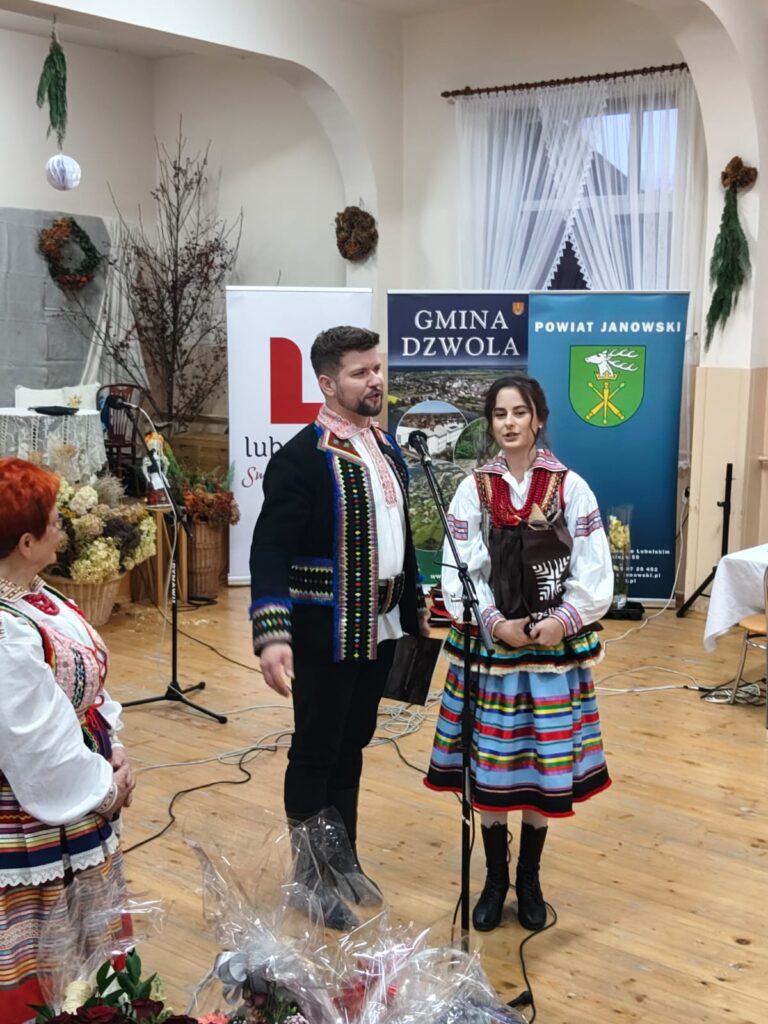 Trzy osoby w kolorowych ludowych strojach, mężczyzna i kobieta przy mikrofonie, tło z banerami i dekoracjami, drewniana podłoga, jasne wnętrze.