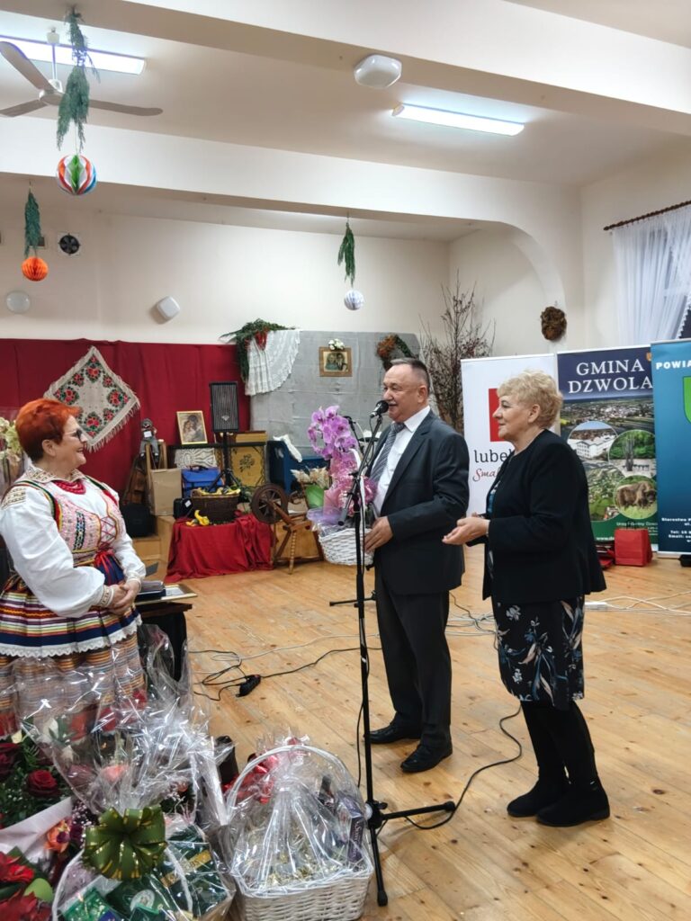 Trzy osoby w sali, mężczyzna w garniturze trzyma bukiet różowych kwiatów przy mikrofonie, kobieta w czarnej sukience i starsza kobieta w kolorowym stroju ludowym, drewniana podłoga, dekoracje.