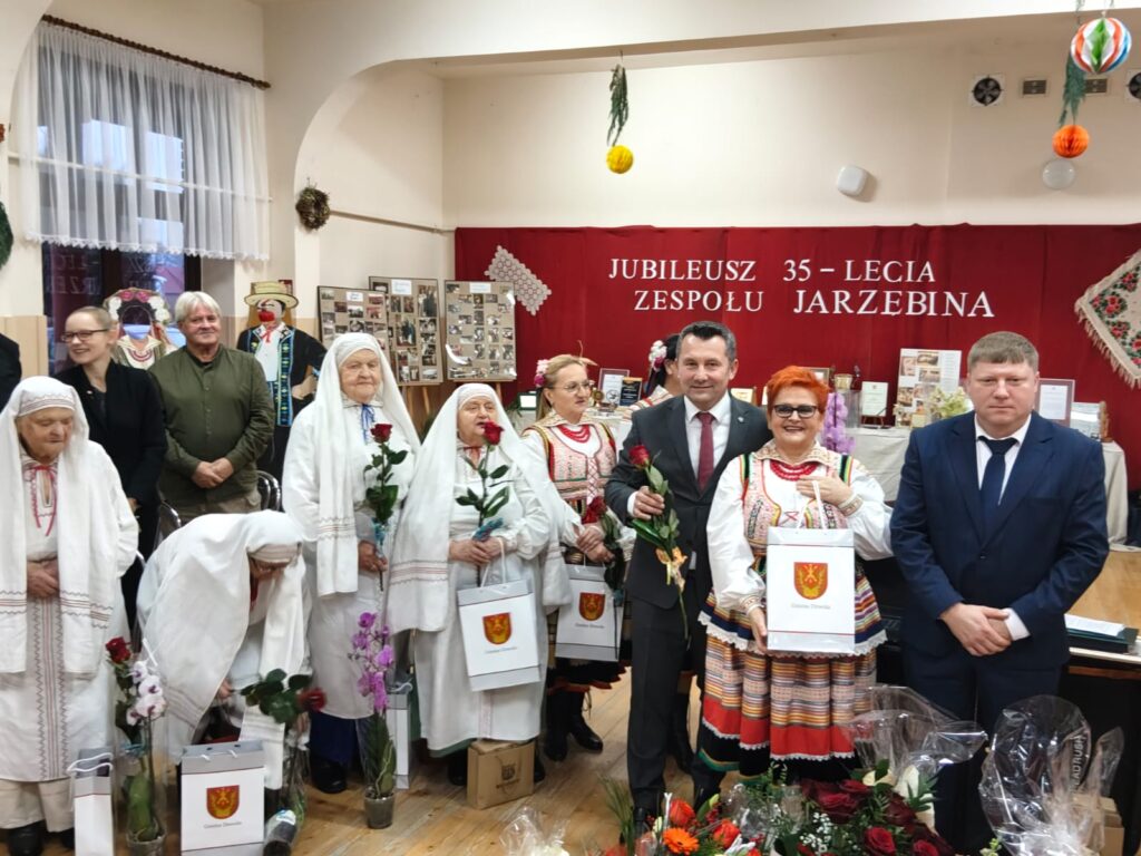 Grupa osób w tradycyjnych i współczesnych strojach na tle czerwonej ściany z napisem jubileusz 35-lecia zespołu Jarzębina, trzymająca róże i torby z herbem.