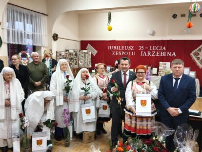 Grupa osób w tradycyjnych i współczesnych strojach na tle czerwonej ściany z napisem jubileusz 35-lecia zespołu Jarzębina, trzymająca róże i torby z herbem.