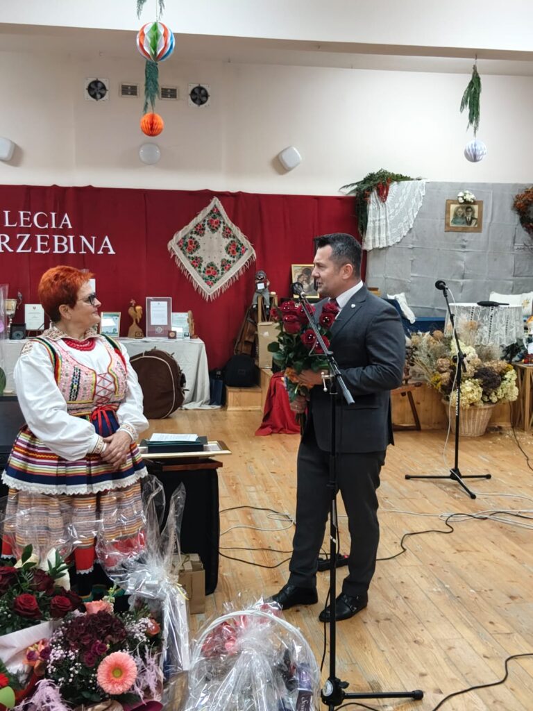 Mężczyzna w garniturze trzyma bukiet czerwonych róż, stoi naprzeciw kobiety w kolorowym stroju ludowym, w tle dekoracje i czerwony napis na ścianie, drewniana podłoga.