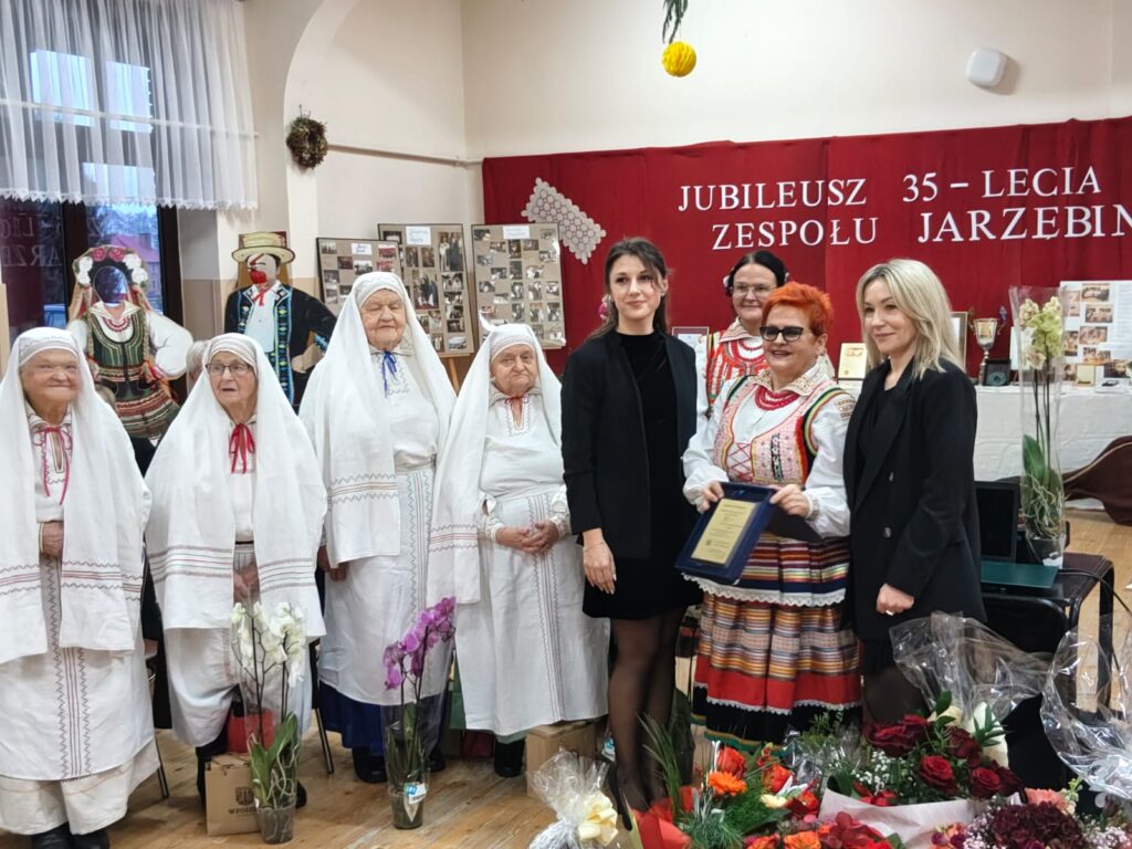 Grupa kobiet, część w białych tradycyjnych strojach ludowych z chustami na głowach, inne w czarnych ubraniach i kolorowym stroju ludowym, na tle czerwonego baneru z napisem.