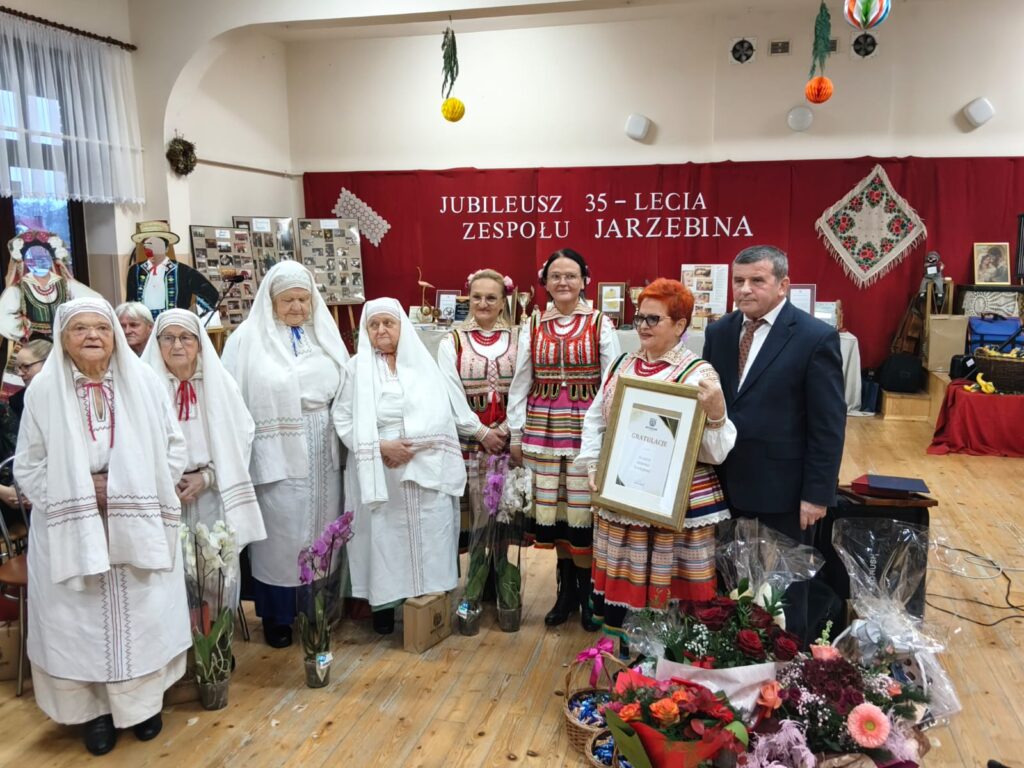 Grupa osób w tradycyjnych strojach ludowych stoi na drewnianej podłodze, w tle czerwony baner z napisem jubileuszowym i dekoracje kwiatowe oraz ramka z gratulacjami.