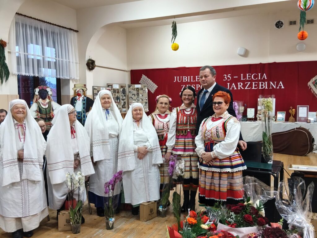 Grupa osób w tradycyjnych strojach ludowych, kobiety w białych chustach i kolorowych haftowanych sukienkach, mężczyzna w garniturze, kwiaty i dekoracje w sali.