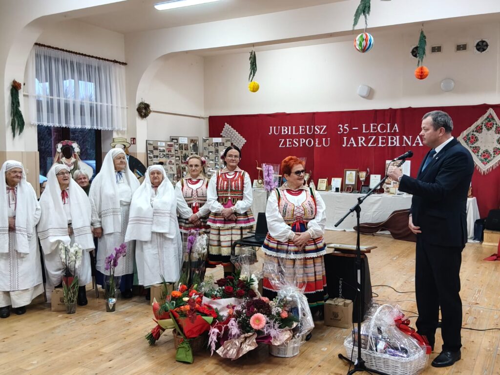 Grupa kobiet w tradycyjnych strojach ludowych, mężczyzna w garniturze przemawia do mikrofonu, kwiaty i kosze prezentów na drewnianej podłodze, czerwony baner z napisem w tle.