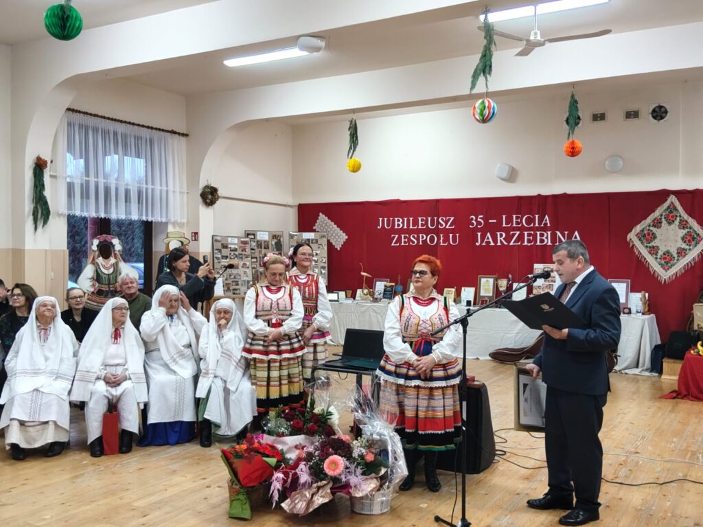 Grupa osób w tradycyjnych strojach ludowych i białych szatach, mężczyzna w garniturze czytający z teczki, dekoracje wiszące, czerwony napis jubileuszowy na ścianie.