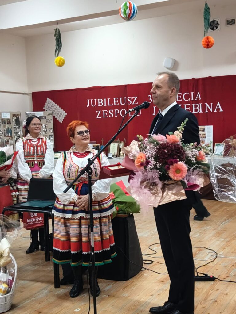 Mężczyzna w garniturze z bukietem kwiatów przy mikrofonie, obok kobiety w kolorowych tradycyjnych strojach ludowych na tle czerwonej kurtyny z napisem jubileuszu.