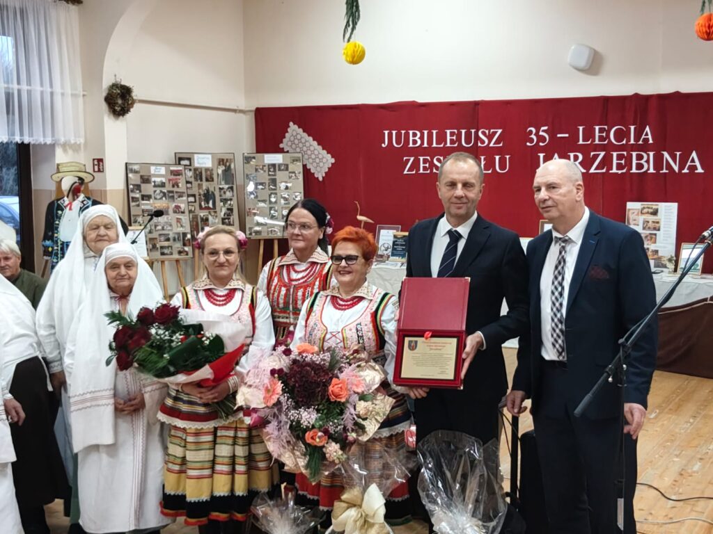 Grupa osób w tradycyjnych strojach ludowych i garniturach stoi na tle czerwonego baneru z napisem jubileusz 35-lecia zespołu Jarzębina, kwiaty i pamiątkowa tablica.