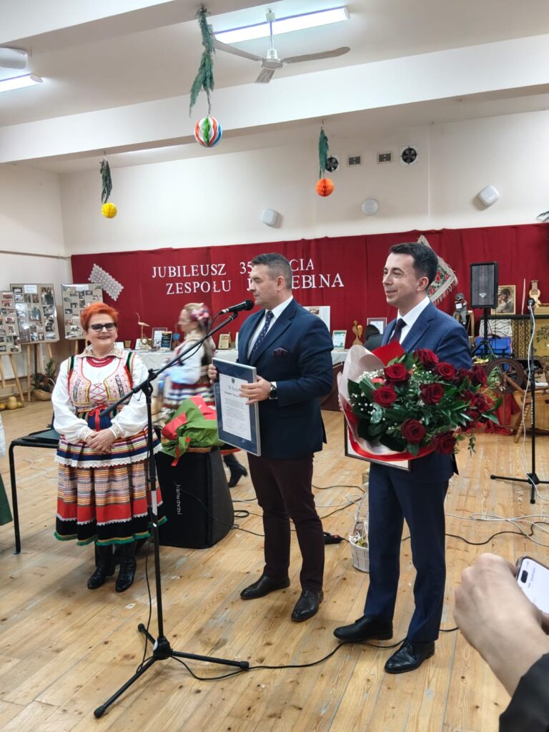 Trzy osoby stoją na drewnianej podłodze, dwie w garniturach, jedna kobieta w kolorowym stroju ludowym, tło z czerwonym banerem i wiszącymi kolorowymi kulami.