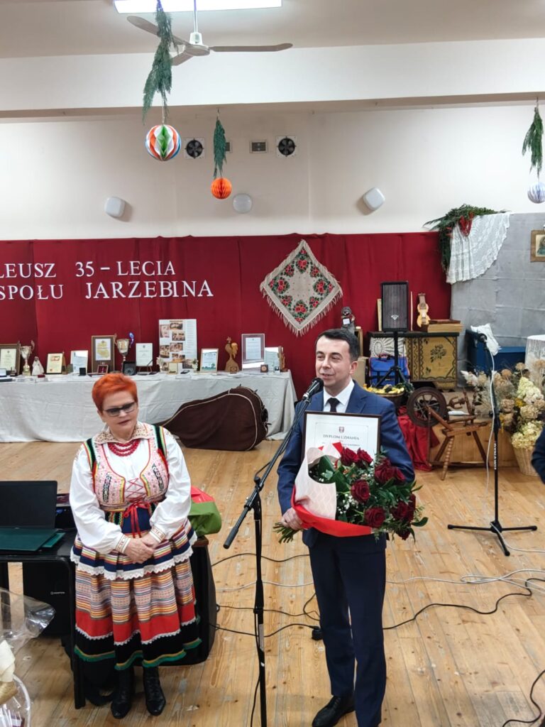 Mężczyzna w garniturze z bukietem czerwonych róż i dyplomem przy mikrofonie, kobieta w kolorowym stroju ludowym obok, tło z czerwonym materiałem i nagrodami na stołach.