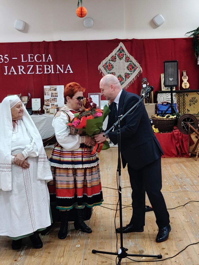 Mężczyzna w garniturze wręcza bukiet kwiatów kobiecie w kolorowym stroju ludowym, obok stoi starsza kobieta w białym tradycyjnym ubraniu, na tle bordowej ściany z napisem.