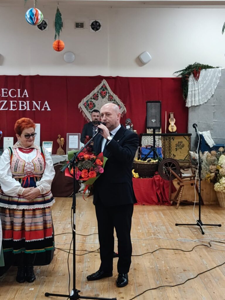 Mężczyzna w czarnym garniturze z bukietem czerwonych i różowych kwiatów mówi do mikrofonu, obok kobieta w kolorowym stroju ludowym, za nimi czerwone tło i dekoracje.